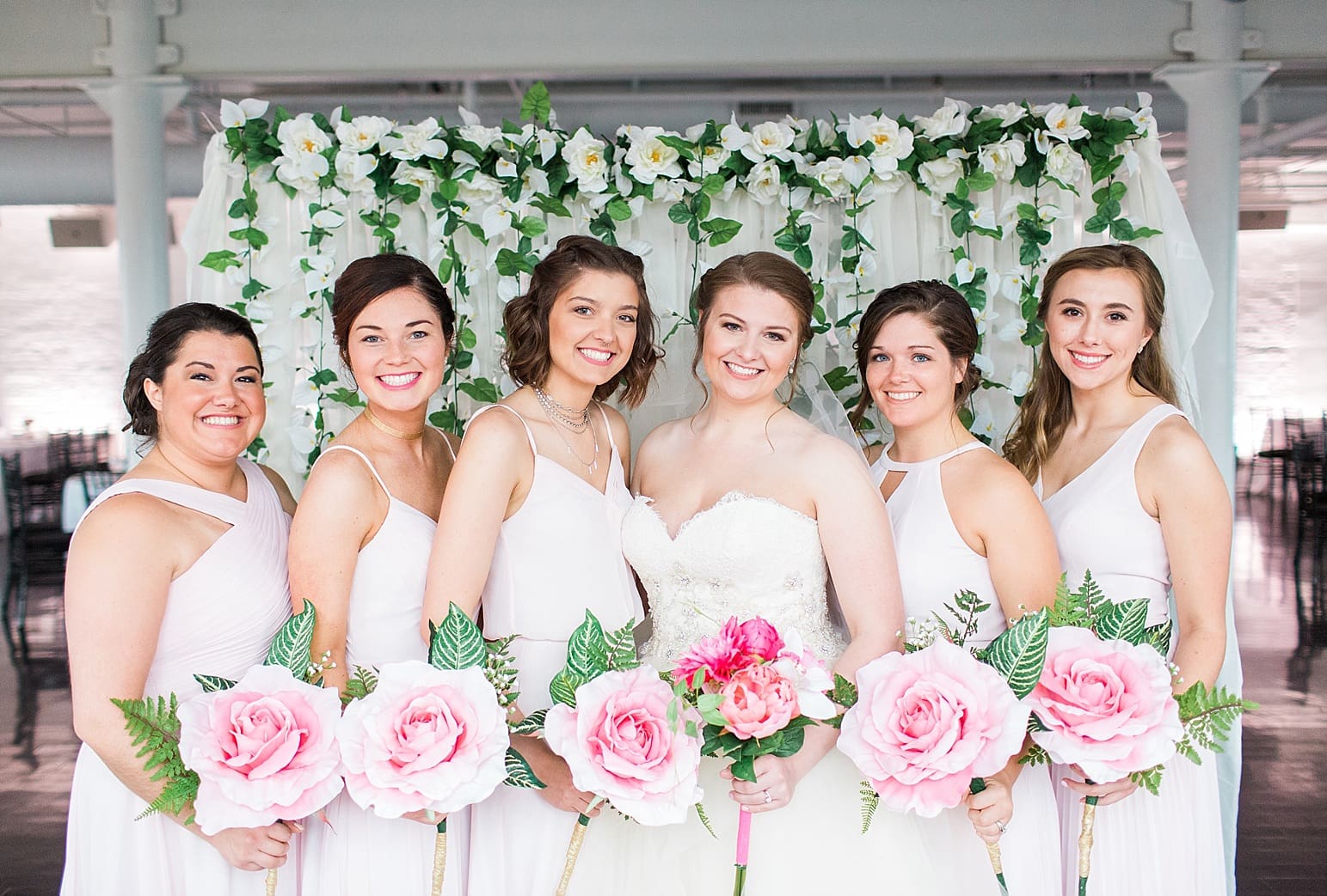 Arielle Peters Photography | Bride and bridesmaids holding bouquets by floral backdrop on wedding day at Loft 310 in Kalamazoo, Michigan.