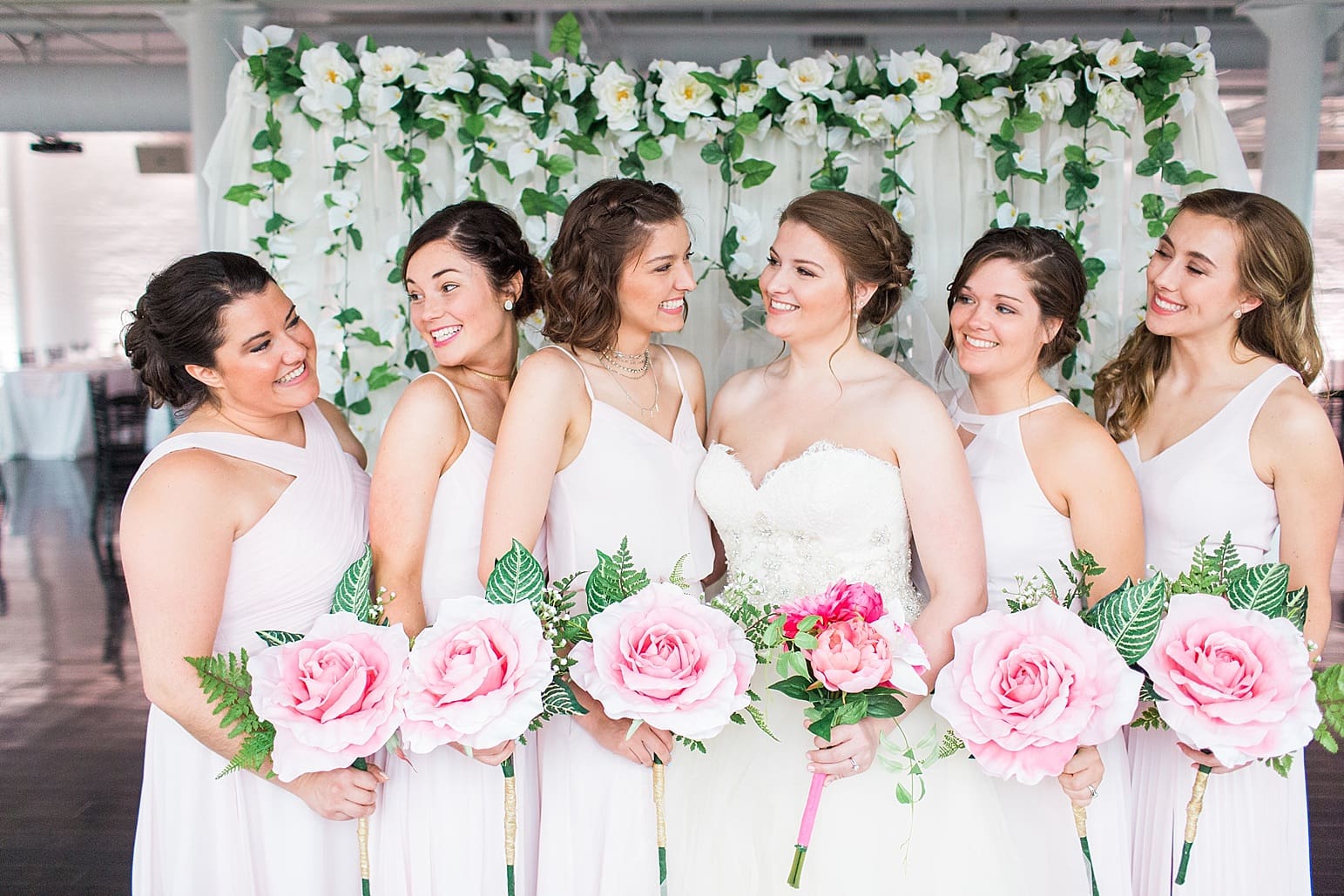 Arielle Peters Photography | Bride and bridesmaids holding bouquets by floral backdrop on wedding day at Loft 310 in Kalamazoo, Michigan.
