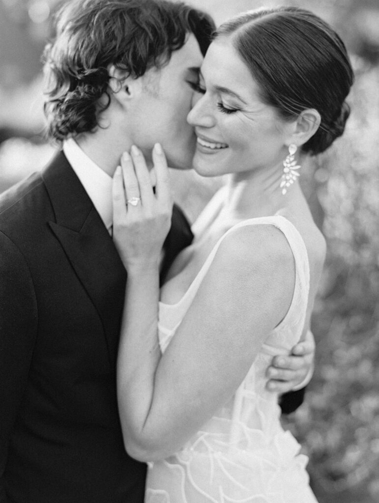 Black and white photo of a groom kissing his brides cheek.