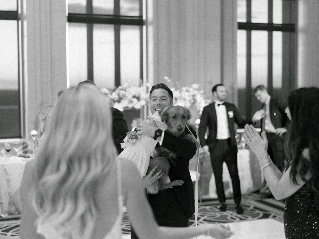 Black and white photo of a groom holding his dog during the wedding reception