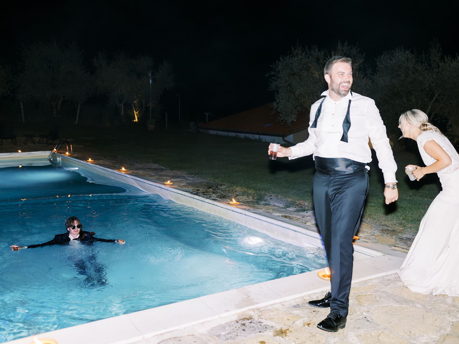 Bride and groom laughing at their wedding guest who jumped into a pool