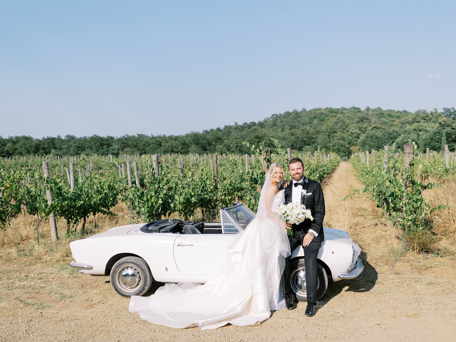 Bride and grooms outdoor summer wedding portraits with a classic white car in the hills of Tuscany, Italy.