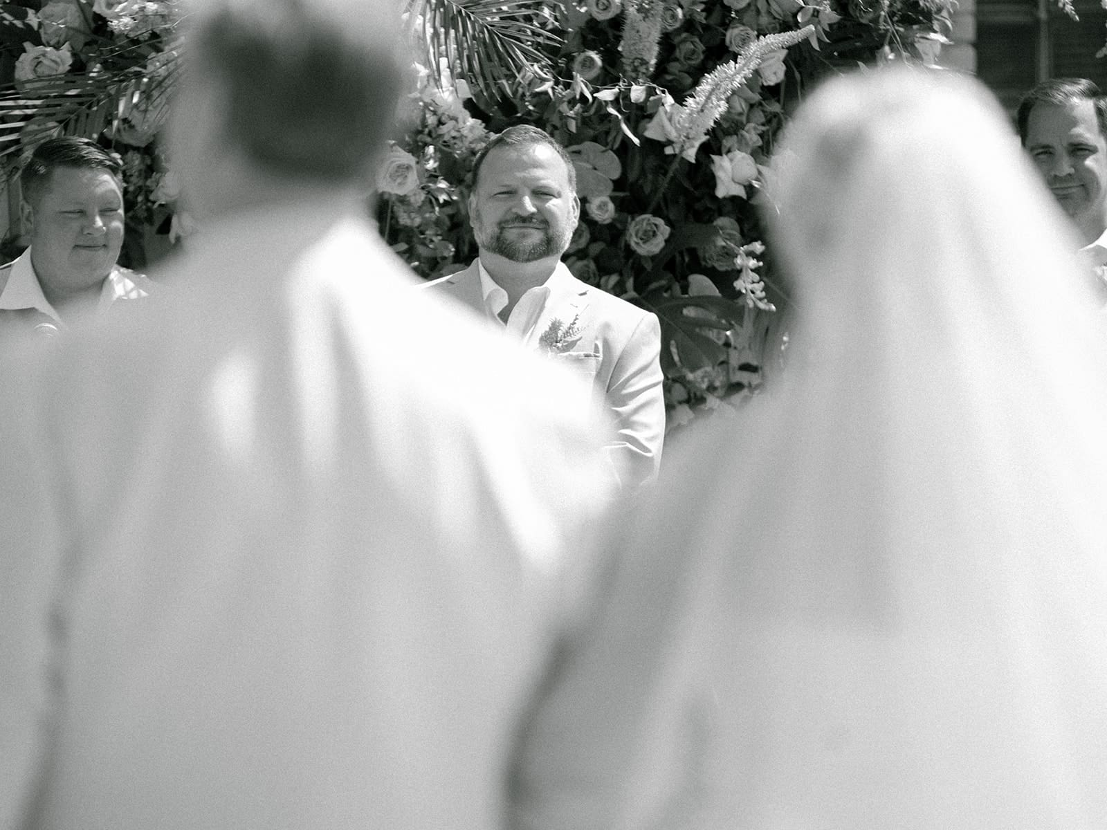 Black and white photo of a groom watching his bride being walked down the aisle by her father during their Journeyman Distillery wedding ceremony in Three Oaks, Michigan