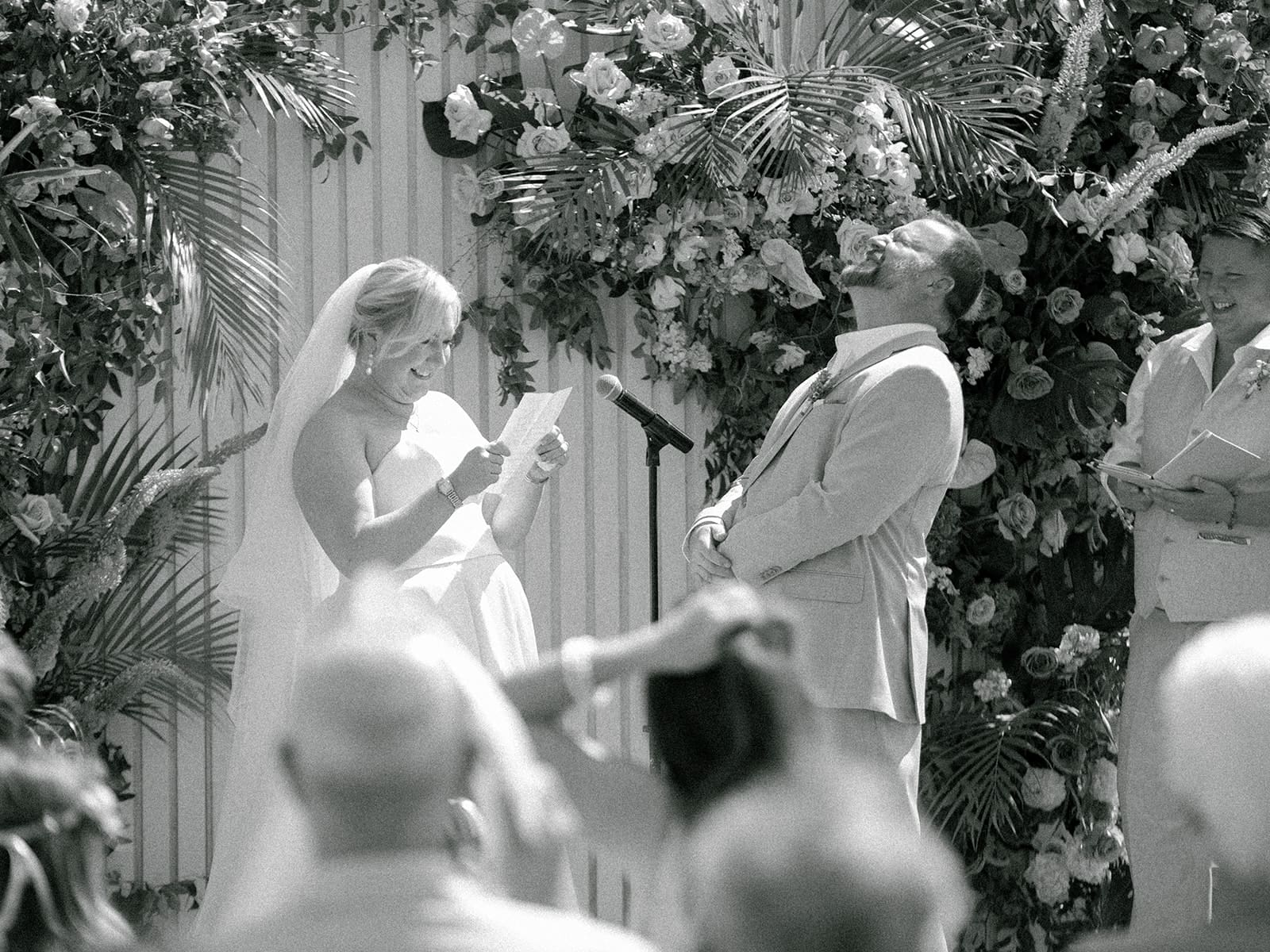 Black and white photo of a groom laughing during while his bride reads her vows to him