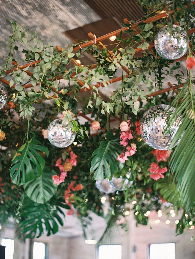 A lush overhead wedding reception floral installation with tropical leaves and disco balls