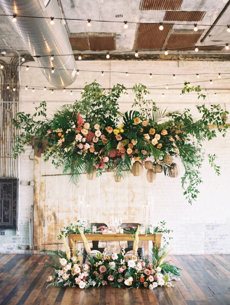 A Journeyman Distillery wedding reception sweethearts table with lush floral arrangements and an overhead installation with hanging wicker lanterns