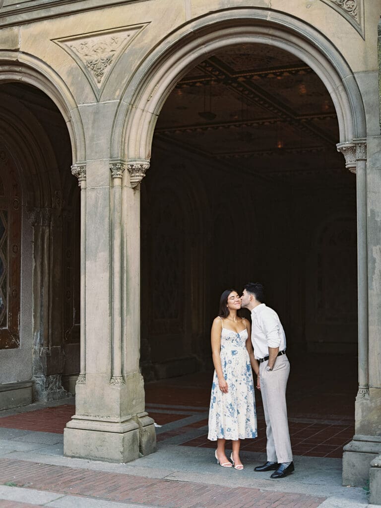 Man kissing his fiancé on the cheek for their Central Park engagement photos at Bethesda Terrace