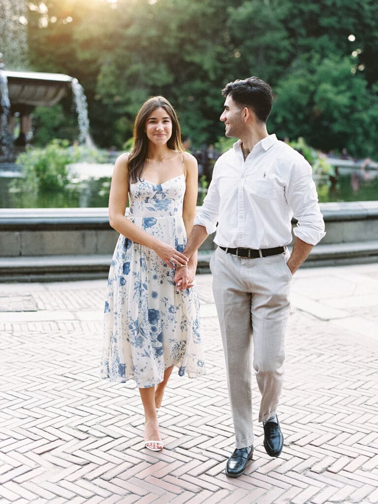 Man and woman walking and holding hands during their Central Park engagement photos at Bethesda Fountain